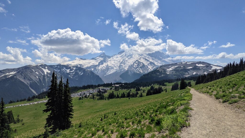 Snow-capped mountains rise above lush green meadows under a bright blue sky with scattered clouds. A winding dirt path leads through the scenery.