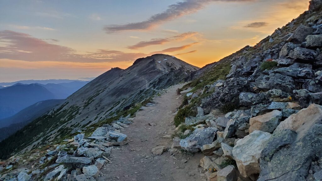 Sunrise over a mountain ridge, highlighting a dirt trail winding alongside rocky outcrops, surrounded by verdant vegetation and distant peaks.