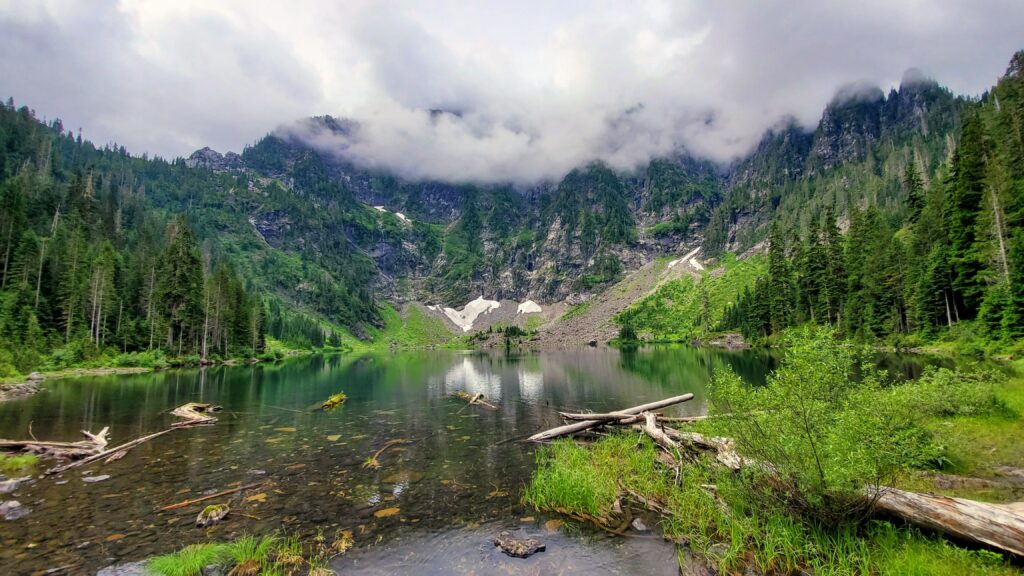 Tranquil lake surrounded by towering mountains and lush forests, with fog shrouding the peaks. Reflective waters enhance the serene landscape.