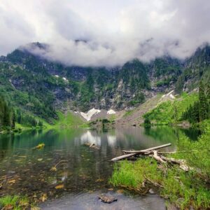 Calm lake reflecting towering mountains under a cloudy sky, surrounded by lush green forest and fallen logs, showcasing stunning natural beauty.