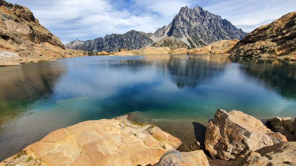 Clear blue lake reflecting rugged mountains under a partly cloudy sky, with a rocky shoreline in the foreground, emphasizing a serene nature setting.