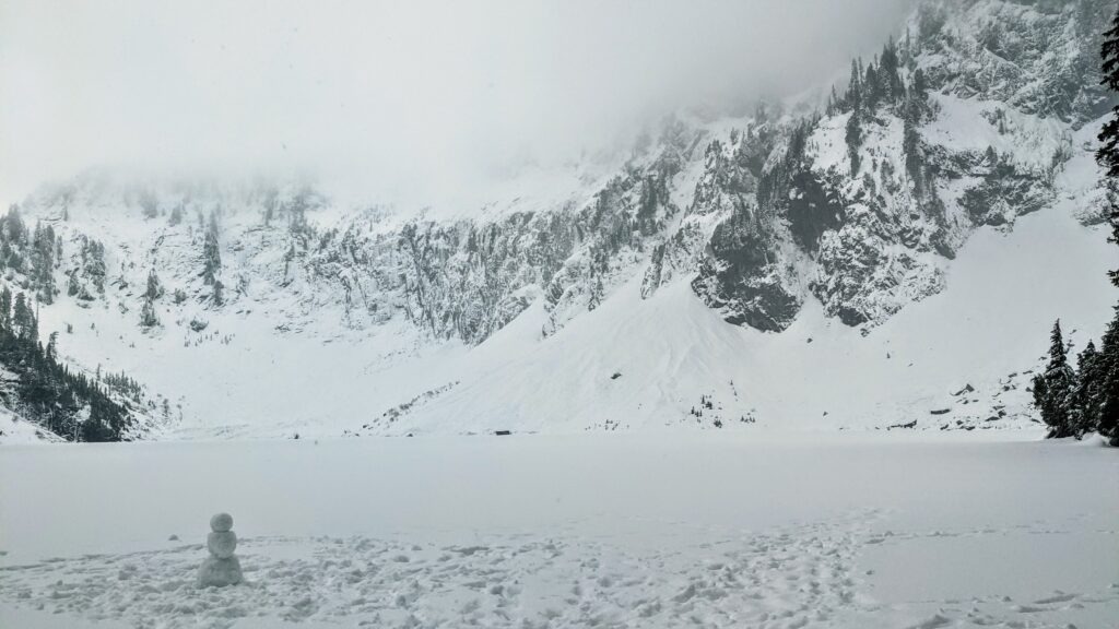 A snowman stands on a snowy shore beside a frozen lake, framed by towering, snow-covered mountains and misty clouds above.