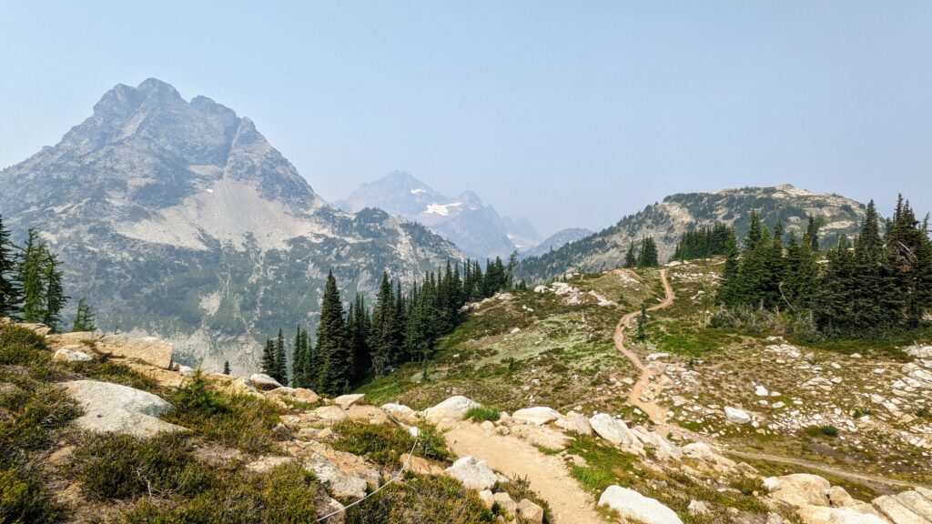 Rocky mountain landscape featuring a winding dirt trail leading through lush greenery and coniferous trees, under a smoky sky.