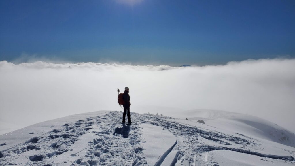 A lone hiker stands on a snowy ridge, overlooking a dense blanket of clouds below, under a clear blue sky.