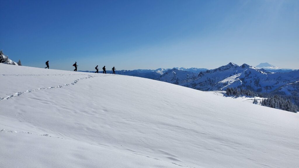 Hikers traverse a snowy landscape, making their way across a white, glistening slope under a clear blue sky, with mountains in the background.