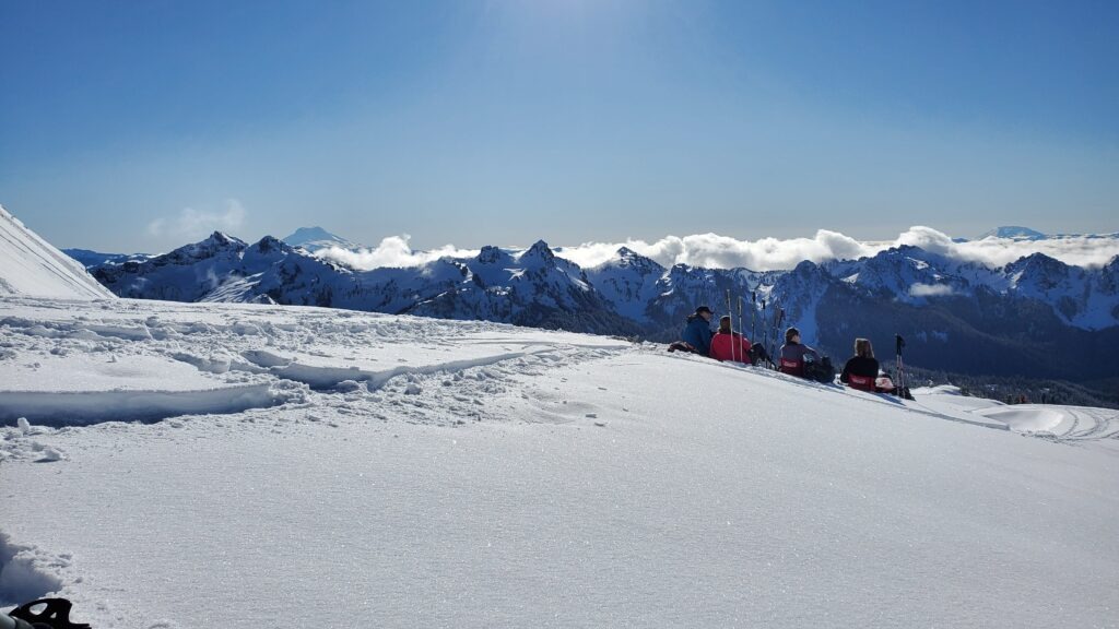 Skiers relax on a snow-covered slope, surrounded by majestic mountains and clear blue skies, enjoying a scenic winter day.