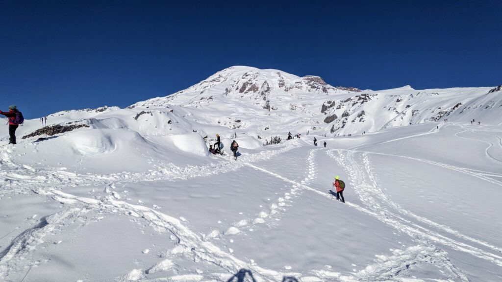 Hikers traverse snowy slopes beneath a clear blue sky, with a prominent mountain peak in the background, showcasing winter adventure.