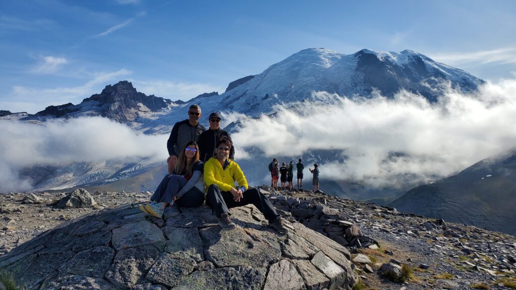 A group of four hikers sitting on a rocky overlook at Second Burroughs Mountain while enjoying guided hikes in Seattle, WA, smiling in front of a snow-capped Mount Rainier.