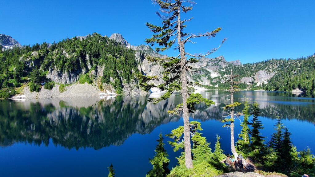 Serene mountain lake reflecting surrounding peaks and evergreen trees. Visitors sit by the water's edge, enjoying the natural beauty and tranquility.