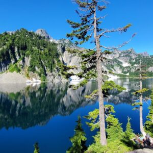 Serene mountain lake reflecting surrounding peaks and evergreen trees. Visitors sit by the water's edge, enjoying the natural beauty and tranquility.